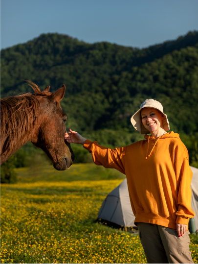 Una donna sorridente in maglione arancione e cappello bianco accarezza affettuosamente un cavallo marrone in un campo fiorito, con tende e colline verdi sullo sfondo